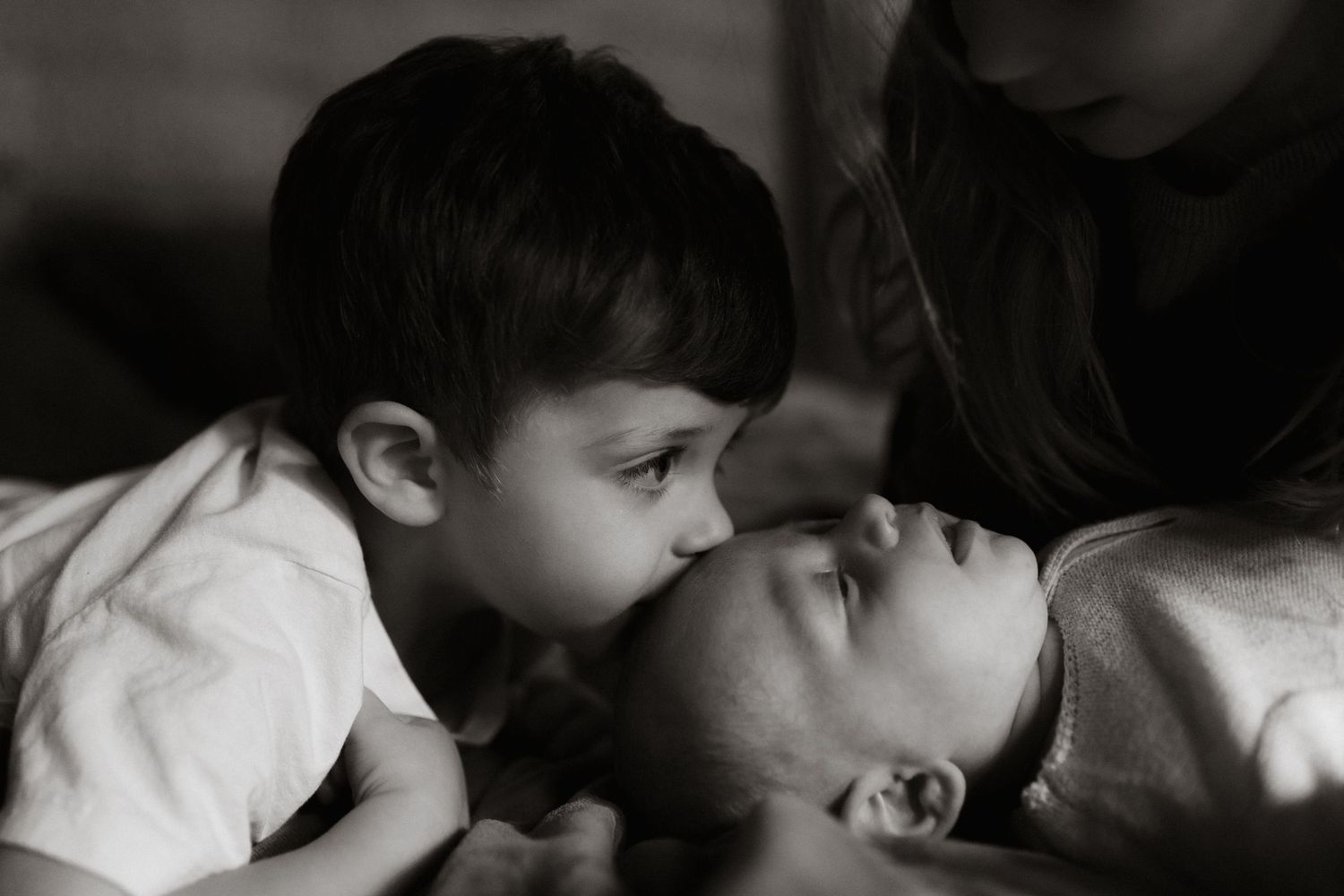 Big brother kissing his new baby sibling's head in black and white