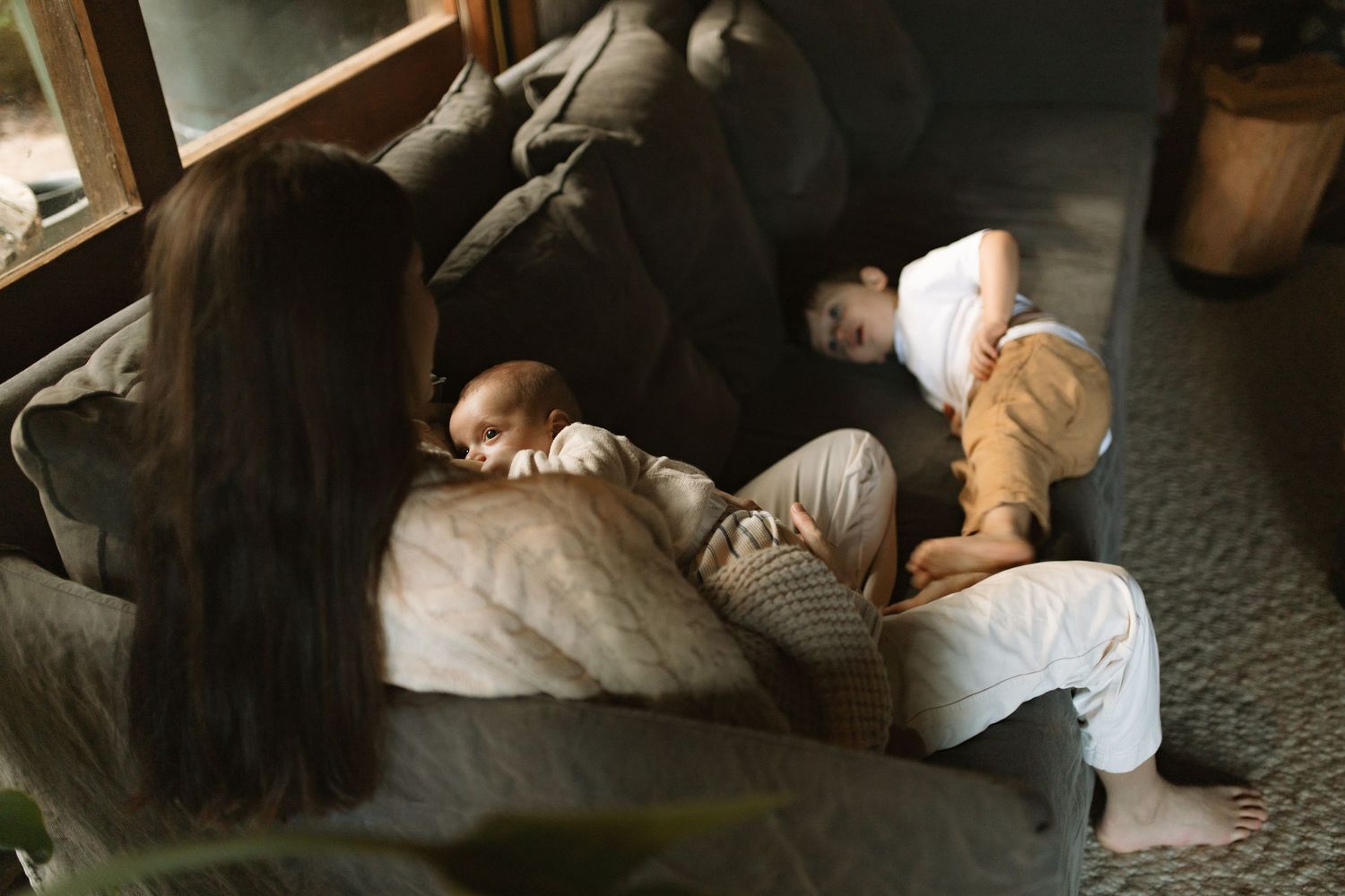 fine art photograph of newborn baby with his mother breastfeeding him while big brother plays on their couch