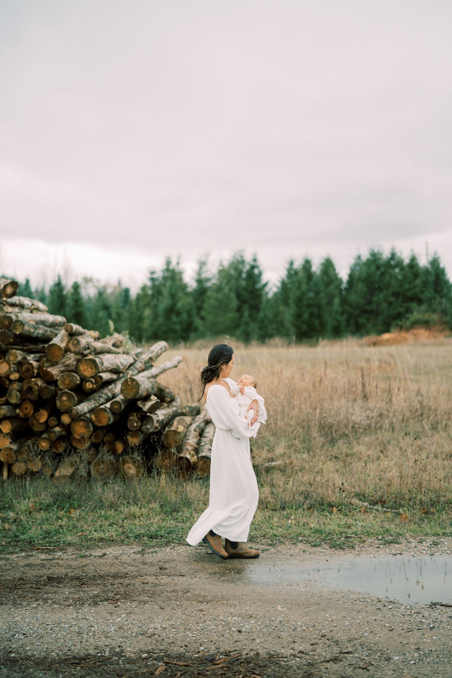 Woman in flowing white dress carries baby while walking past woodpile in misty forest setting.