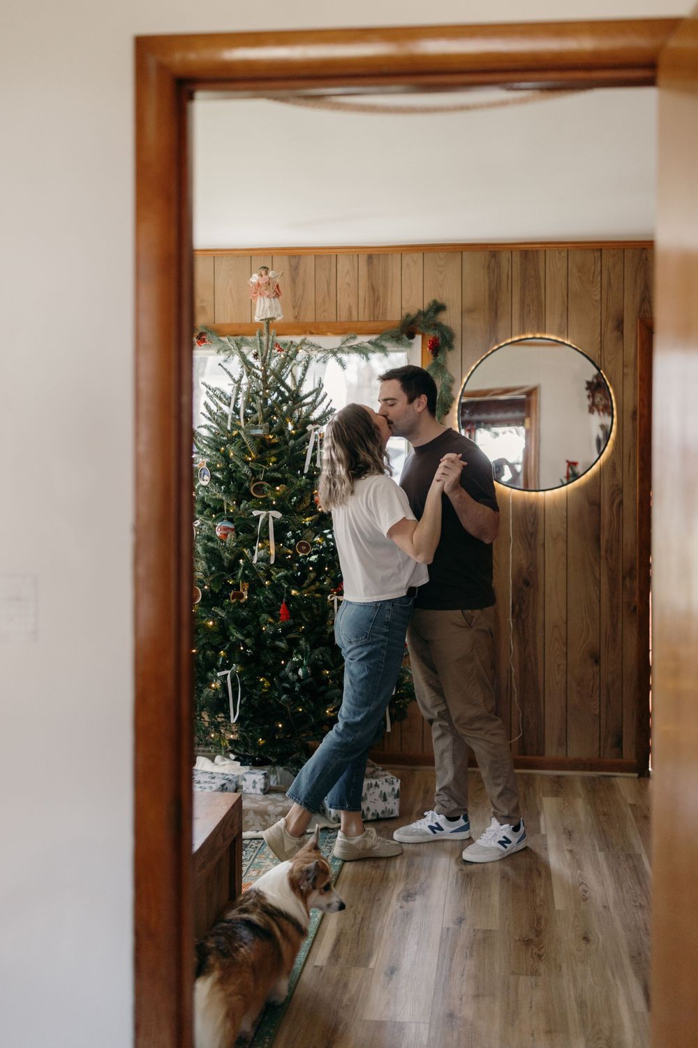 Couple decorating Christmas tree in wood-paneled room with dog watching nearby.