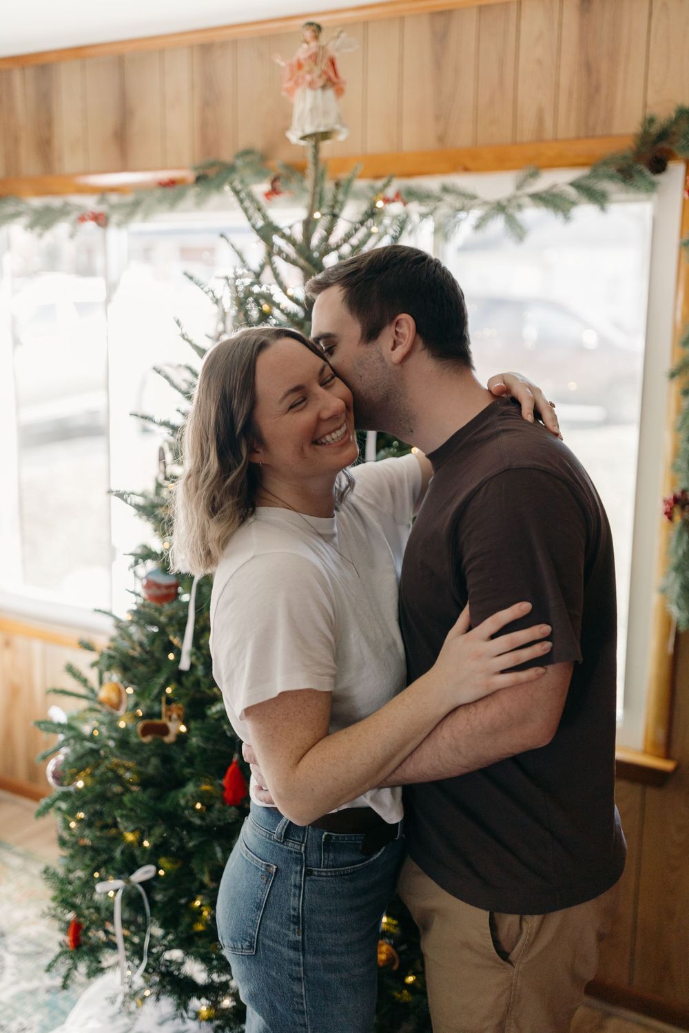A couple shares an intimate moment in front of a decorated Christmas tree with evergreen garlands.