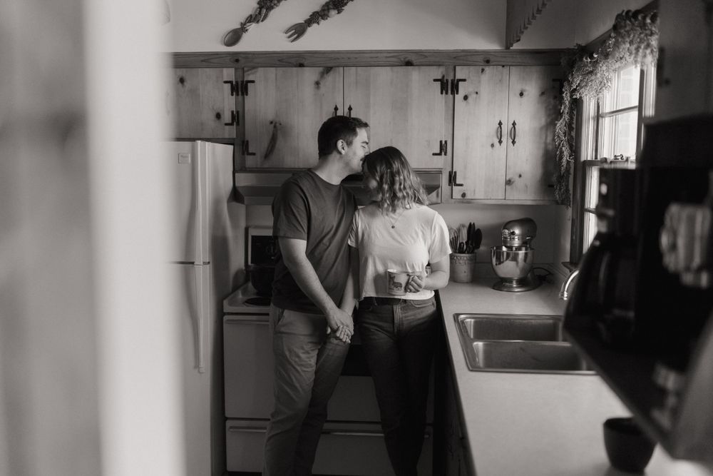 A black and white photograph of a couple sharing a tender moment in their kitchen.