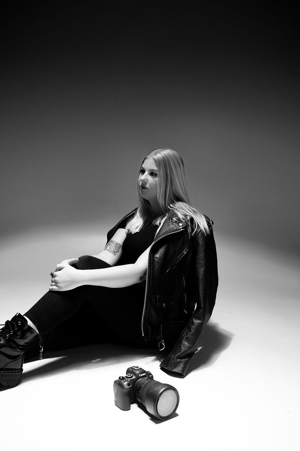 Black and white portrait of someone sitting on floor in leather jacket with camera nearby.