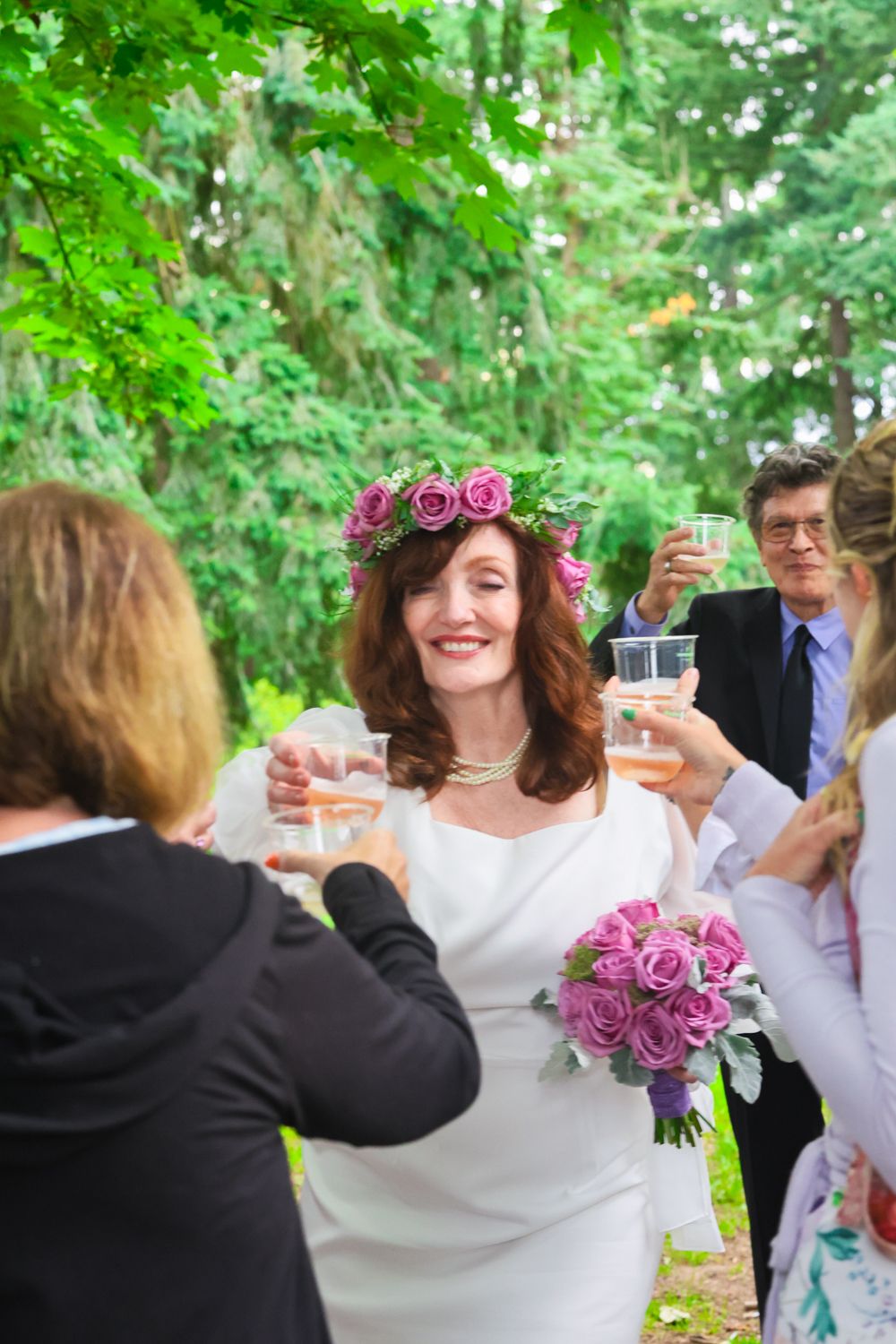 A wedding celebration in a garden setting with people raising glasses for a toast while wearing floral decorations.