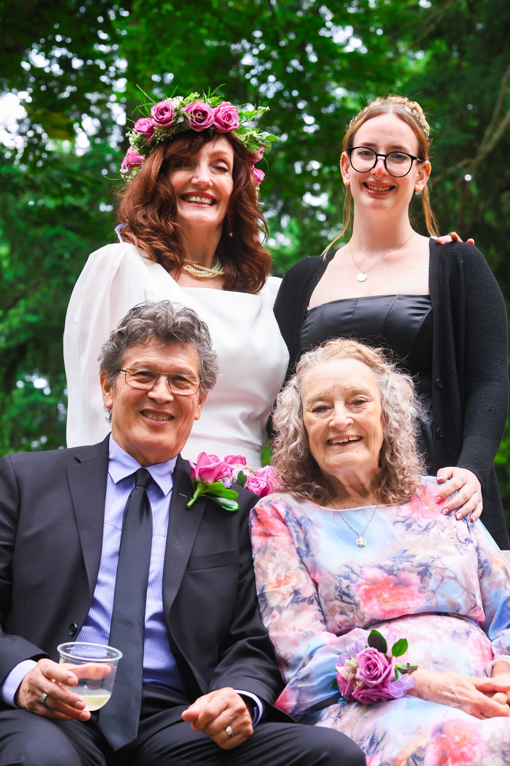 A couple seated outdoors in formal attire with pink roses and greenery in the background.