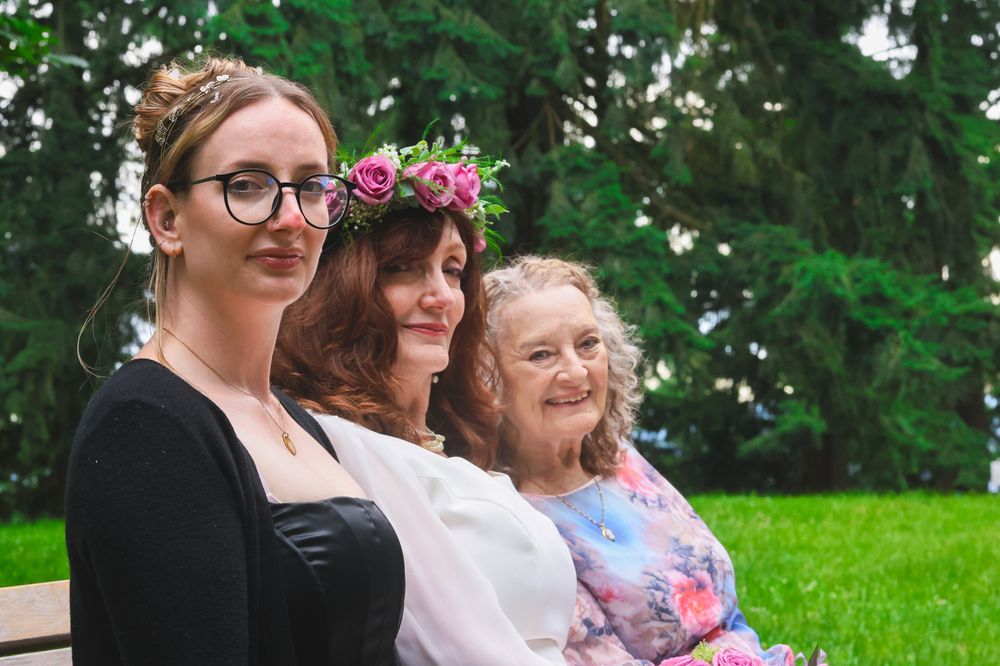 Three people wearing casual attire pose together outdoors in a park with green trees in the background.