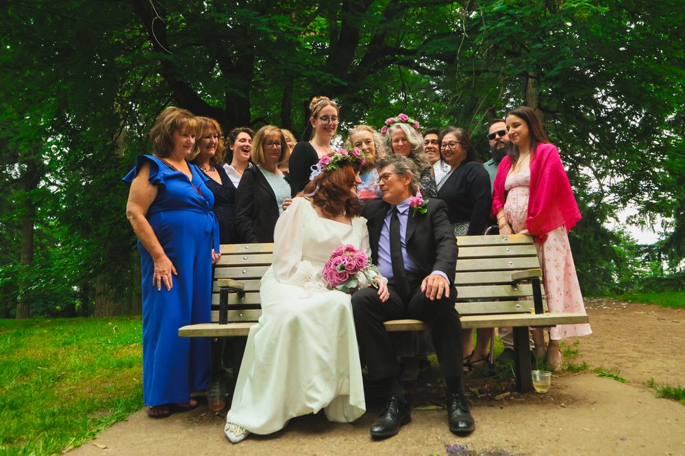 Wedding guests gather on rustic wooden benches surrounded by lush green trees during an outdoor celebration.