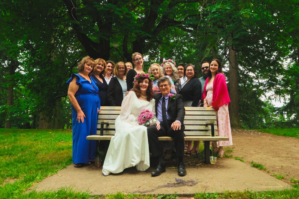 A wedding party gathering poses together on a garden bench surrounded by lush green trees and foliage.