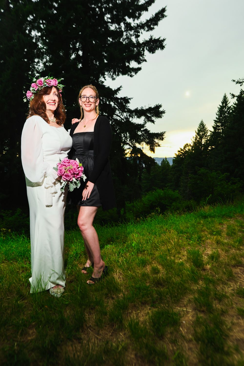 Outdoor hillside photo session at sunset captures two people standing together among evergreen trees.