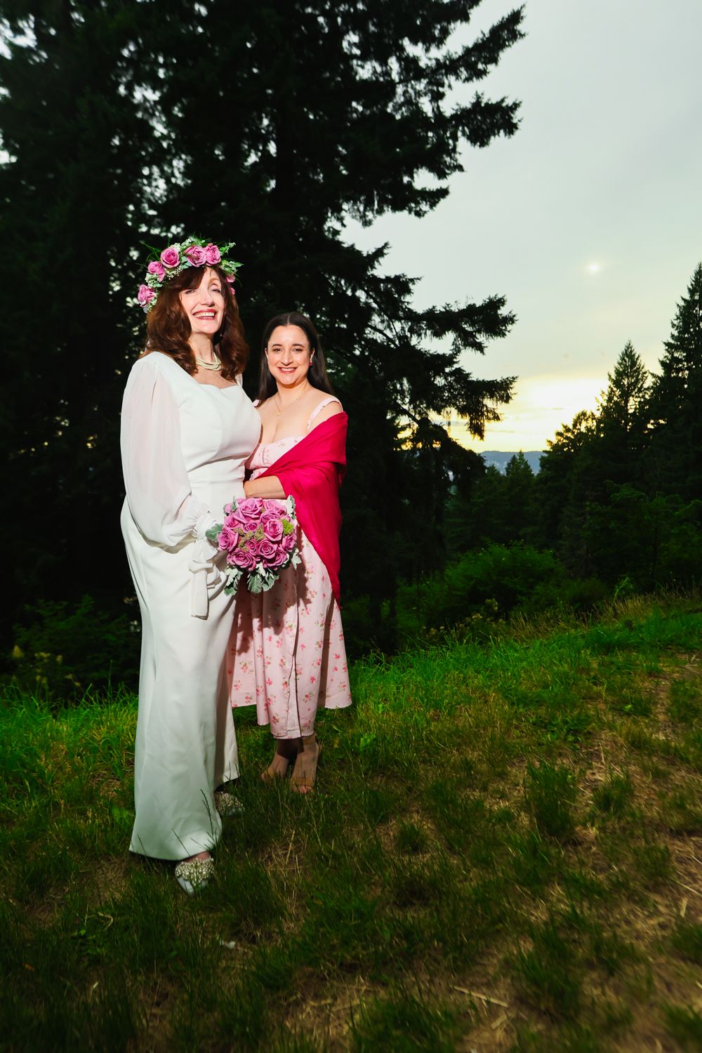 Two people stand together during a wedding photo session in an outdoor field with evergreen trees at sunset.