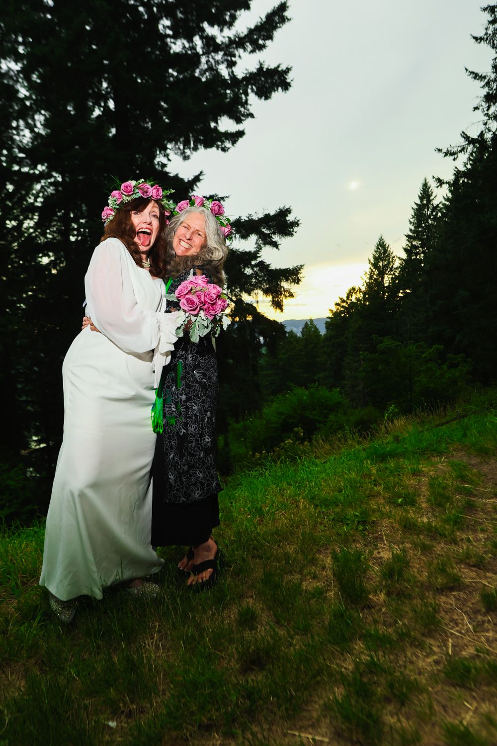 Two people in floral crowns share a joyous moment outdoors at sunset, with evergreen trees and mountains in background.