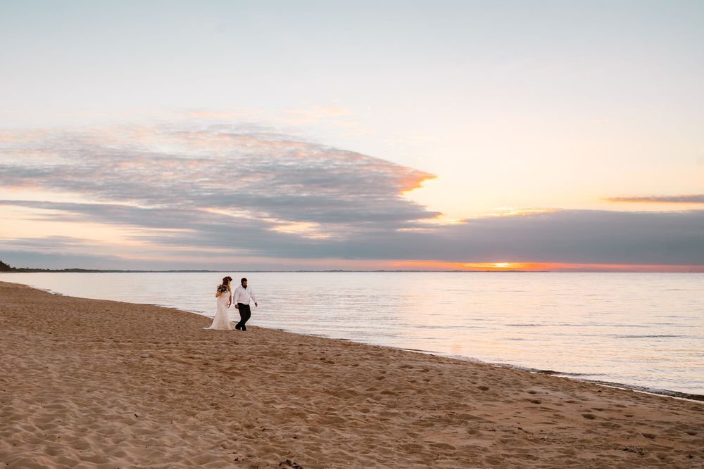 Two figures stand on a sandy beach at sunset, silhouetted against the pink and orange sky over calm waters.
