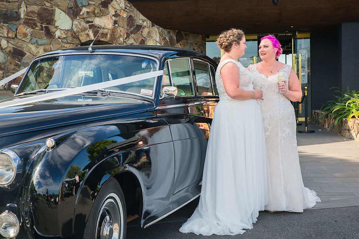Couple in white wedding dresses standing next to a vintage black classic car on their special day.