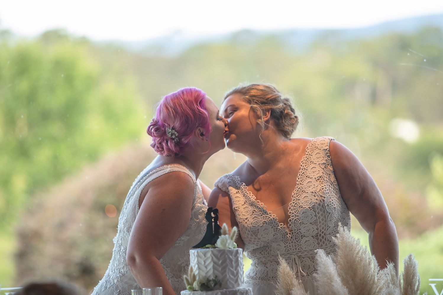A couple shares a romantic sunset kiss at their outdoor wedding surrounded by greenery and natural light.