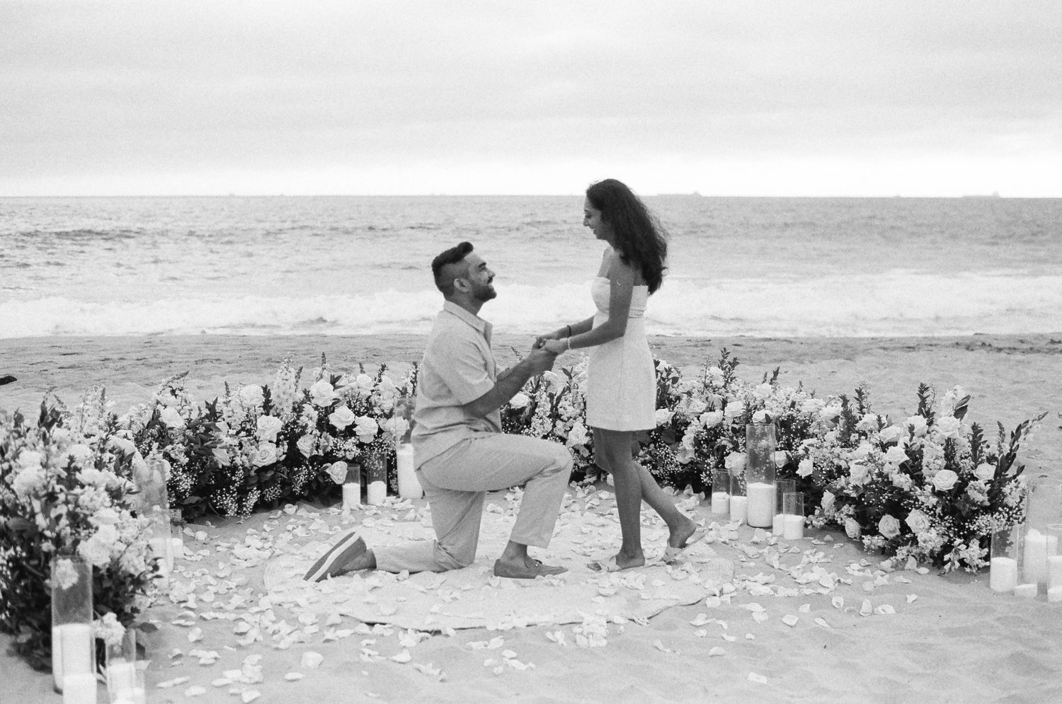 A romantic beach proposal setup with a floral circle arch and candles along the oceanfront at sunset.
