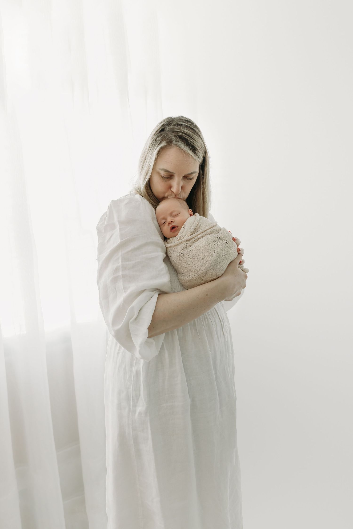 A mother in white clothing tenderly embraces her newborn baby in a bright newborn studio in Tampa