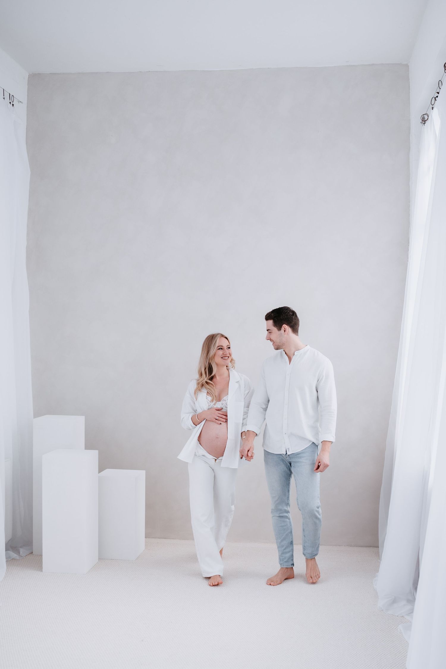 A couple in white outfits share an intimate moment in a bright minimalist room with white decor and pedestals.