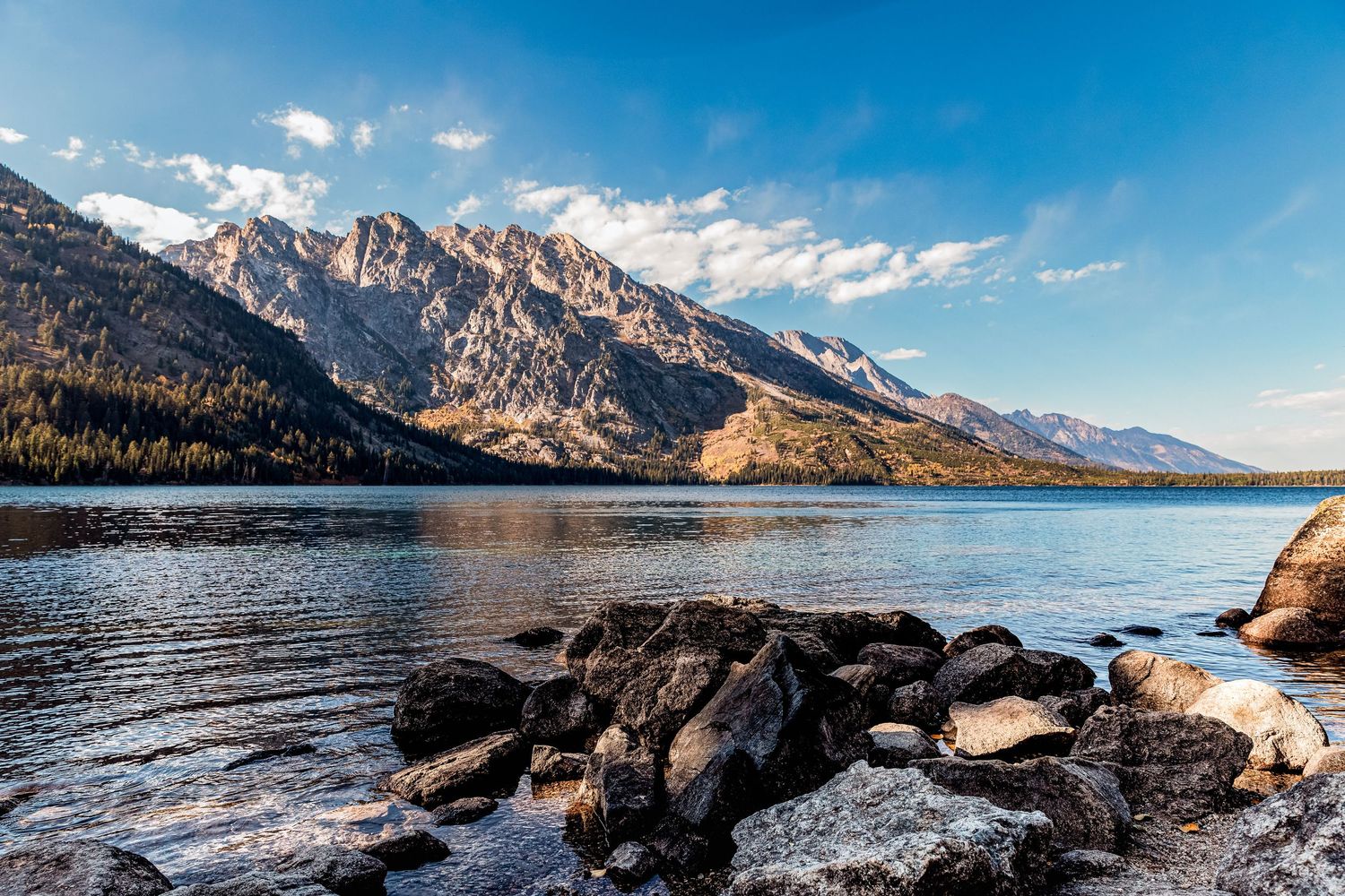 Rugged mountains rise above Jenny Lake with dark rocks in foreground at Grand Teton National Park.