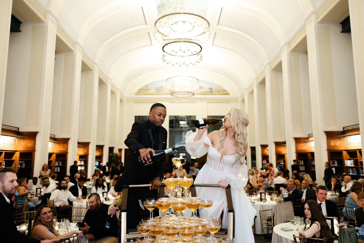 Wedding reception guests enjoy dinner and conversation in an elegant ballroom with arched ceilings and warm lighting.