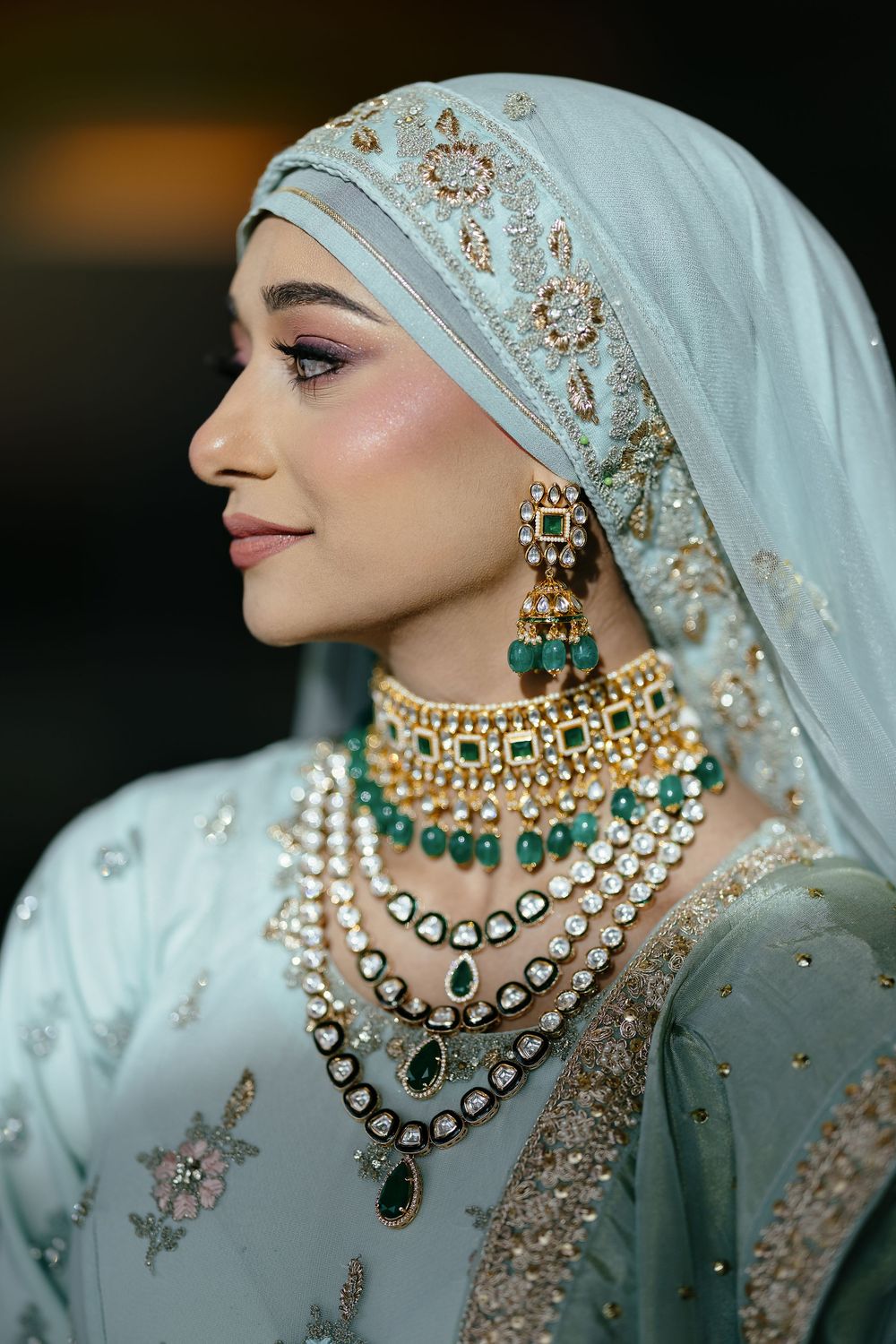 Bride wearing light blue embroidered hijab and layered statement necklaces with emerald and pearl details.