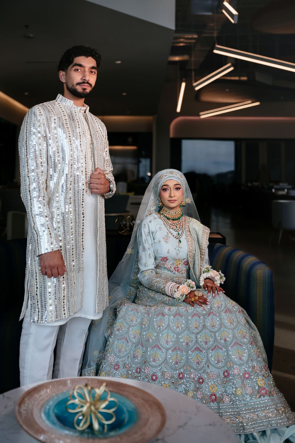 An elegant couple in traditional South Asian wedding attire features embroidered white and pastel designs in a modern setting.