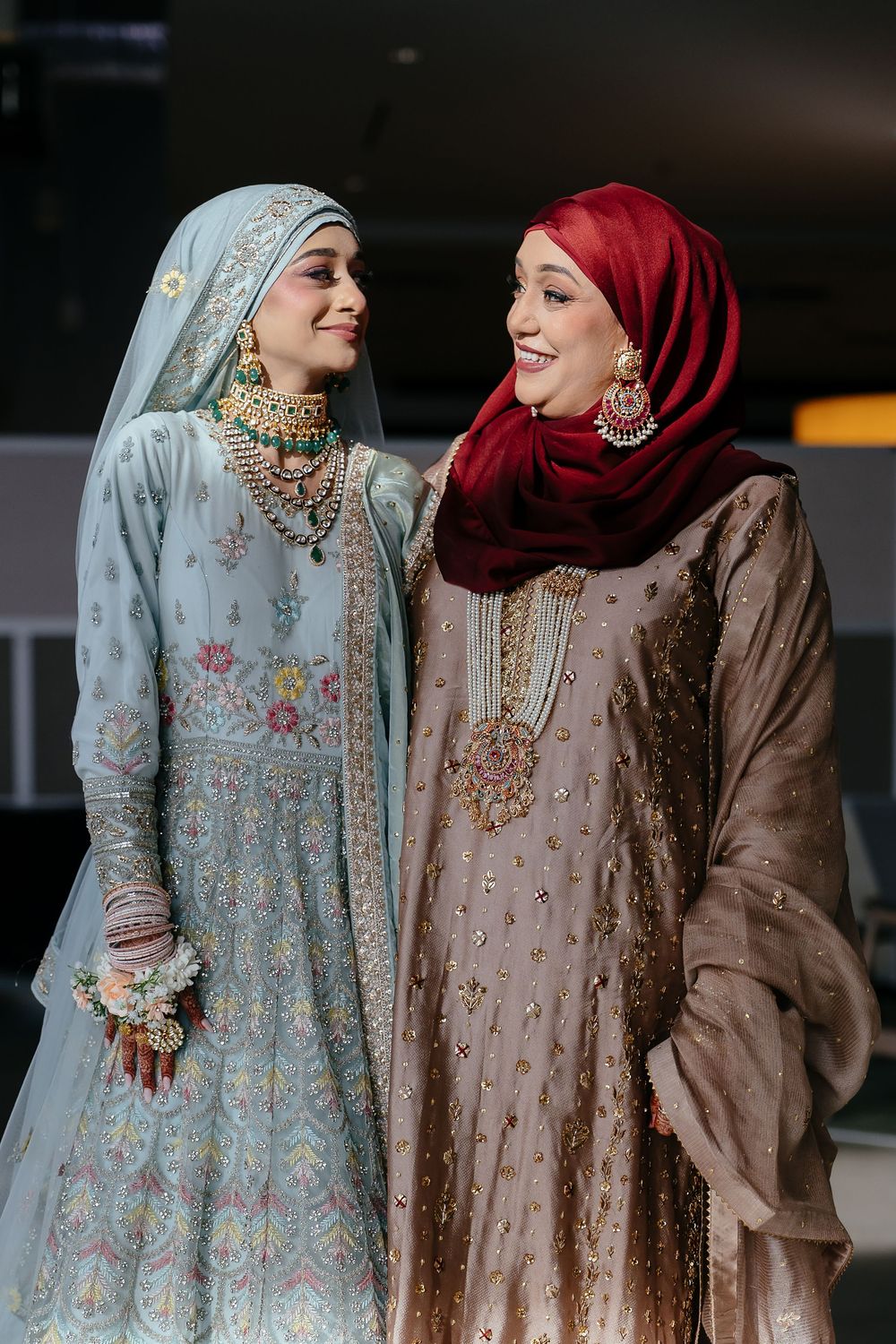 Two people wearing hijabs and traditional formal attire in light blue and burgundy share a joyful moment together.