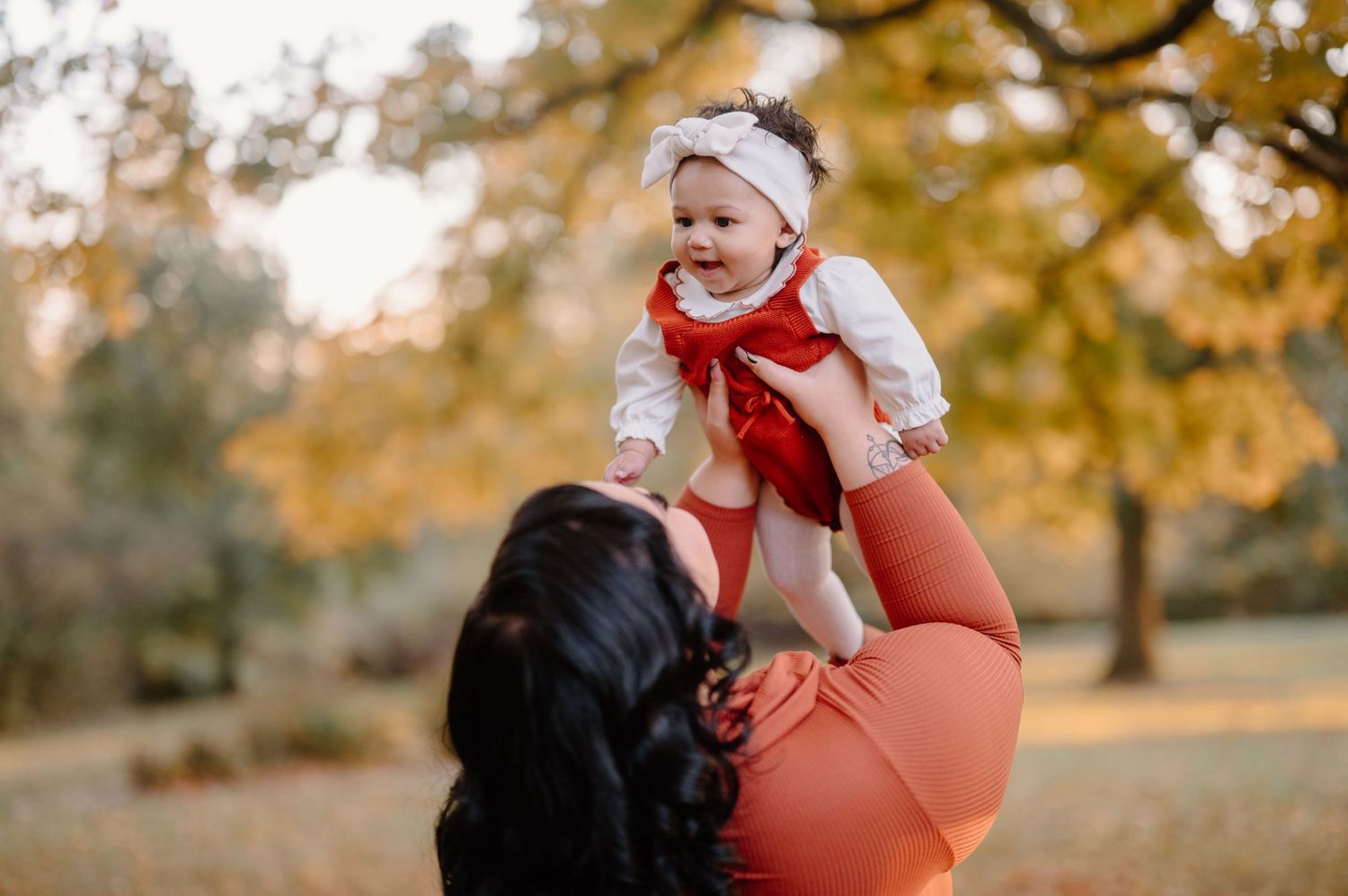 A caregiver lifts a baby wearing a white hat and red scarf during an autumn photo session in a park.