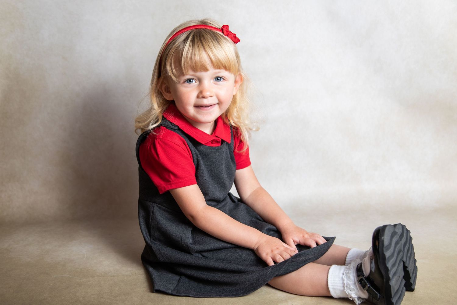 Child seated on gray backdrop wearing red shirt and gray jumper dress with matching hair bow and white socks.