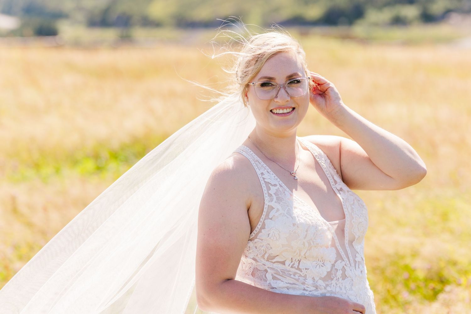 A bride in a white lace wedding dress poses in a sunny golden field while adjusting her flowing veil.