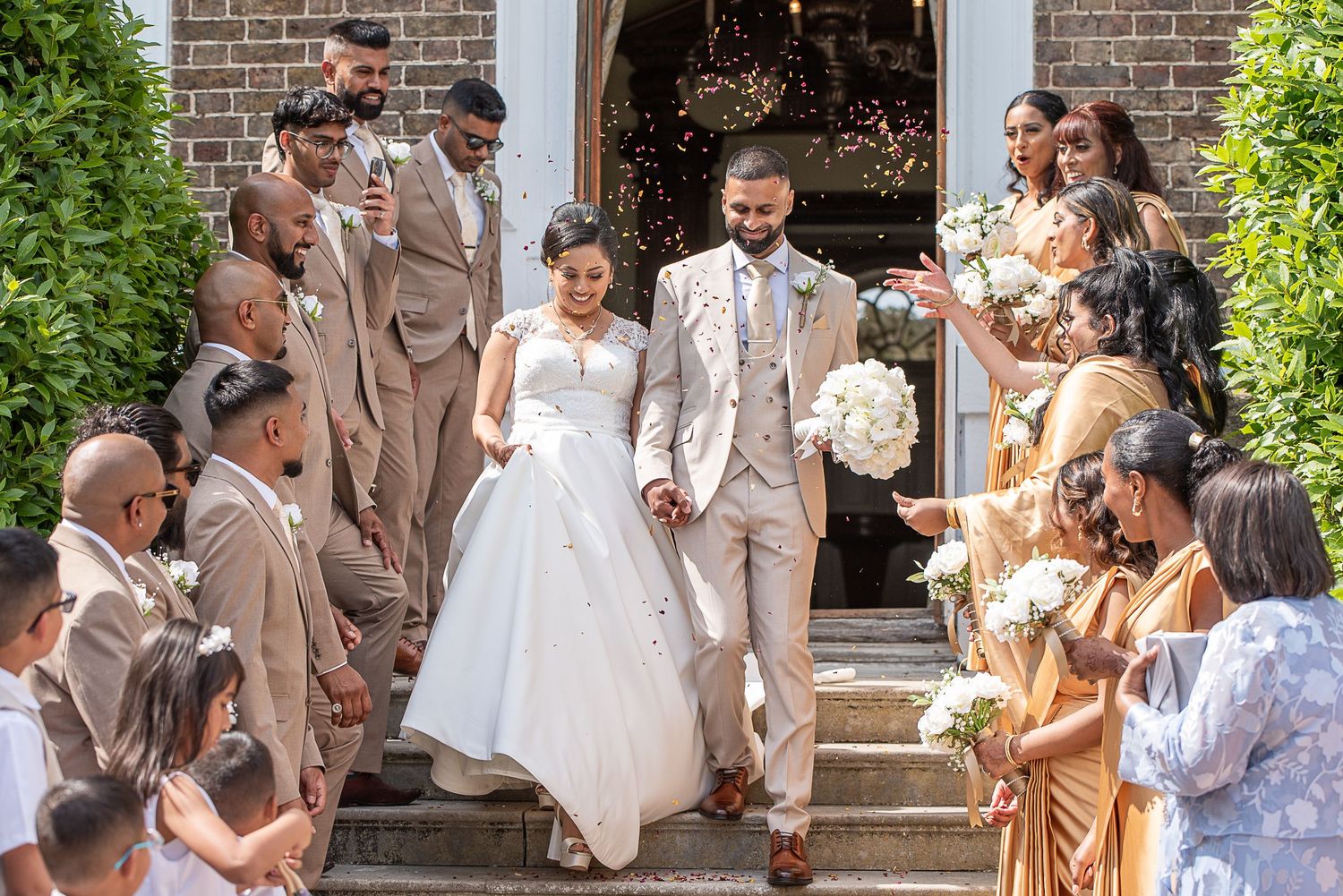Couple come down stairs in sunshine while guests throw petal confetti
