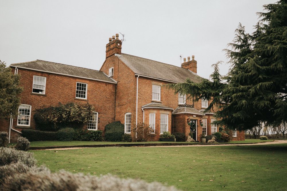 Traditional red brick manor house with white trim windows and multiple chimneys stands behind a manicured lawn.