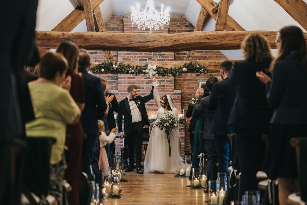 A rustic barn wedding ceremony with wooden beams, elegant chandelier, and guests seated in rows of chairs.
