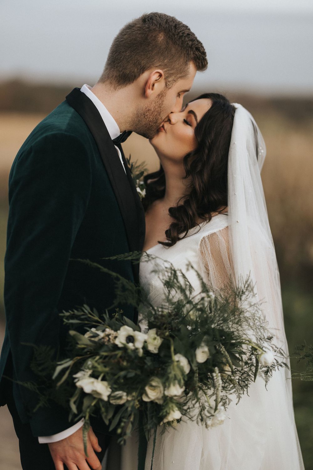 A romantic wedding couple embraces and shares a kiss while holding a beautiful white floral bouquet in an outdoor setting.