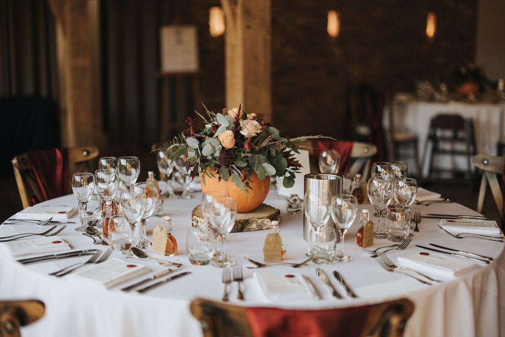 Elegant round wedding reception table with floral centerpiece, white linens, and place settings in rustic venue.