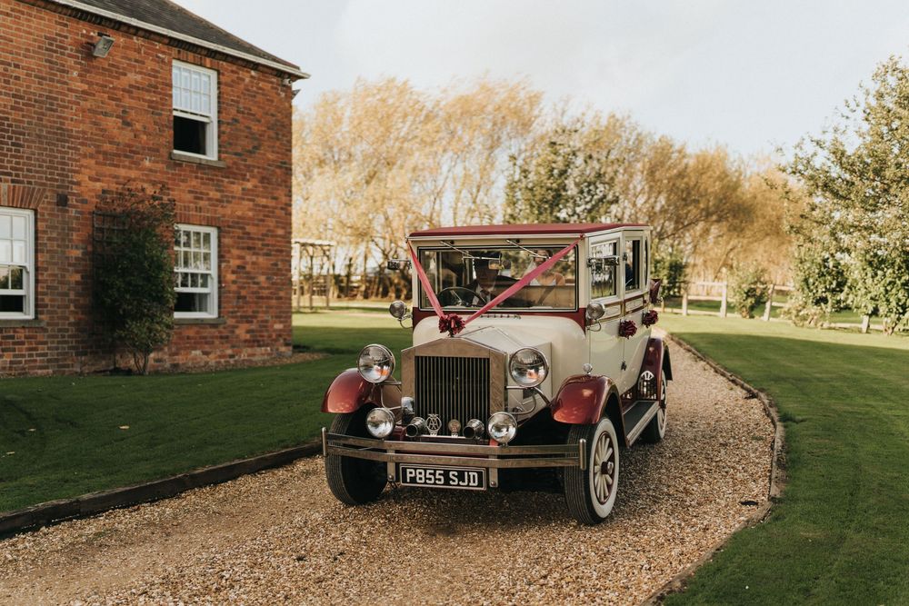 Vintage classic car parked on gravel driveway outside red brick house with autumn trees in background.