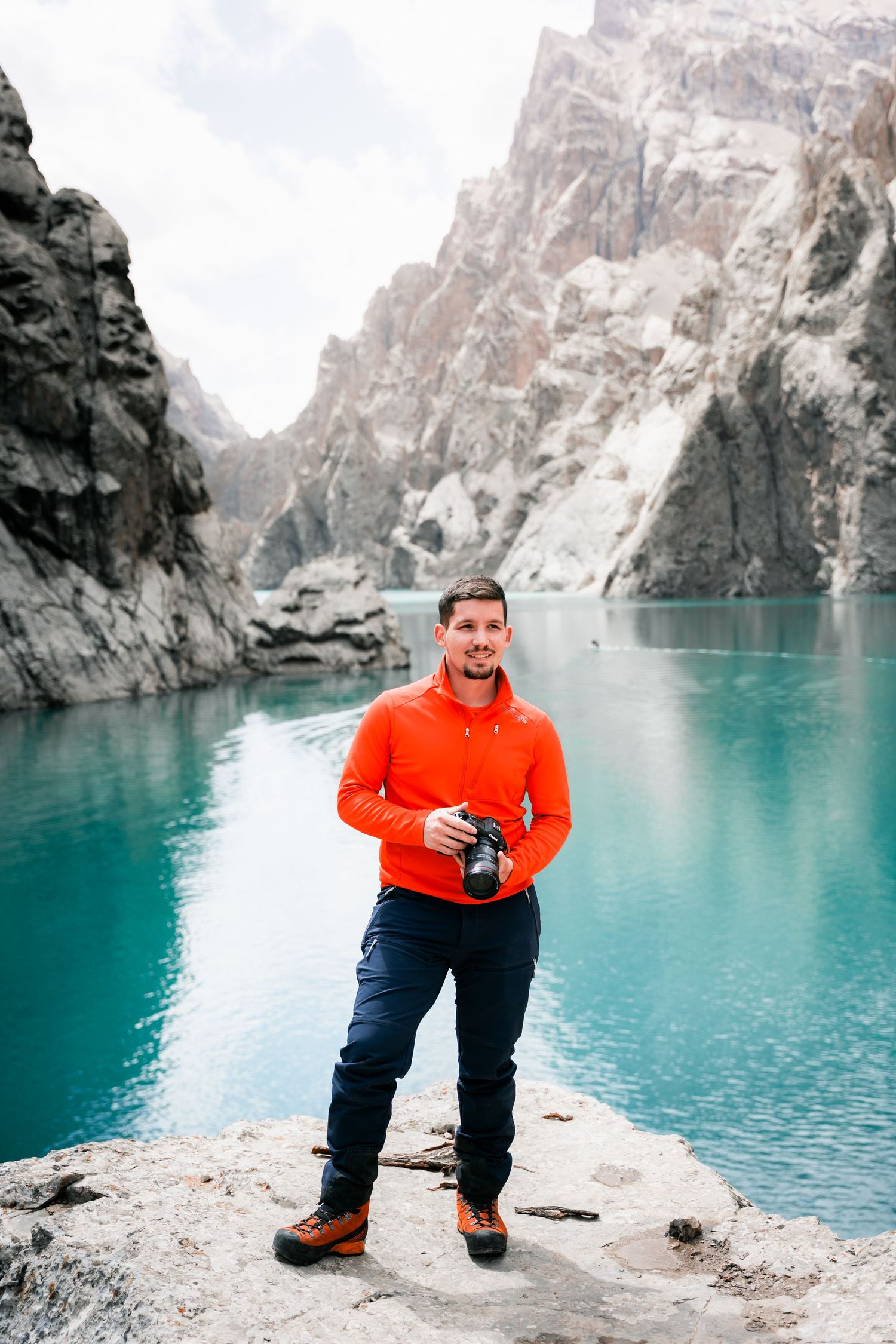 Someone in a bright red sweater stands with a camera at a turquoise alpine lake surrounded by dramatic rocky cliffs.