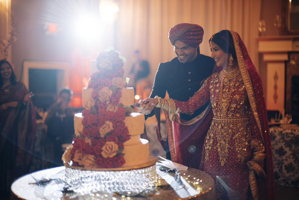 Elegant wedding couple cuts their tiered floral wedding cake at an ornate reception venue with warm lighting.