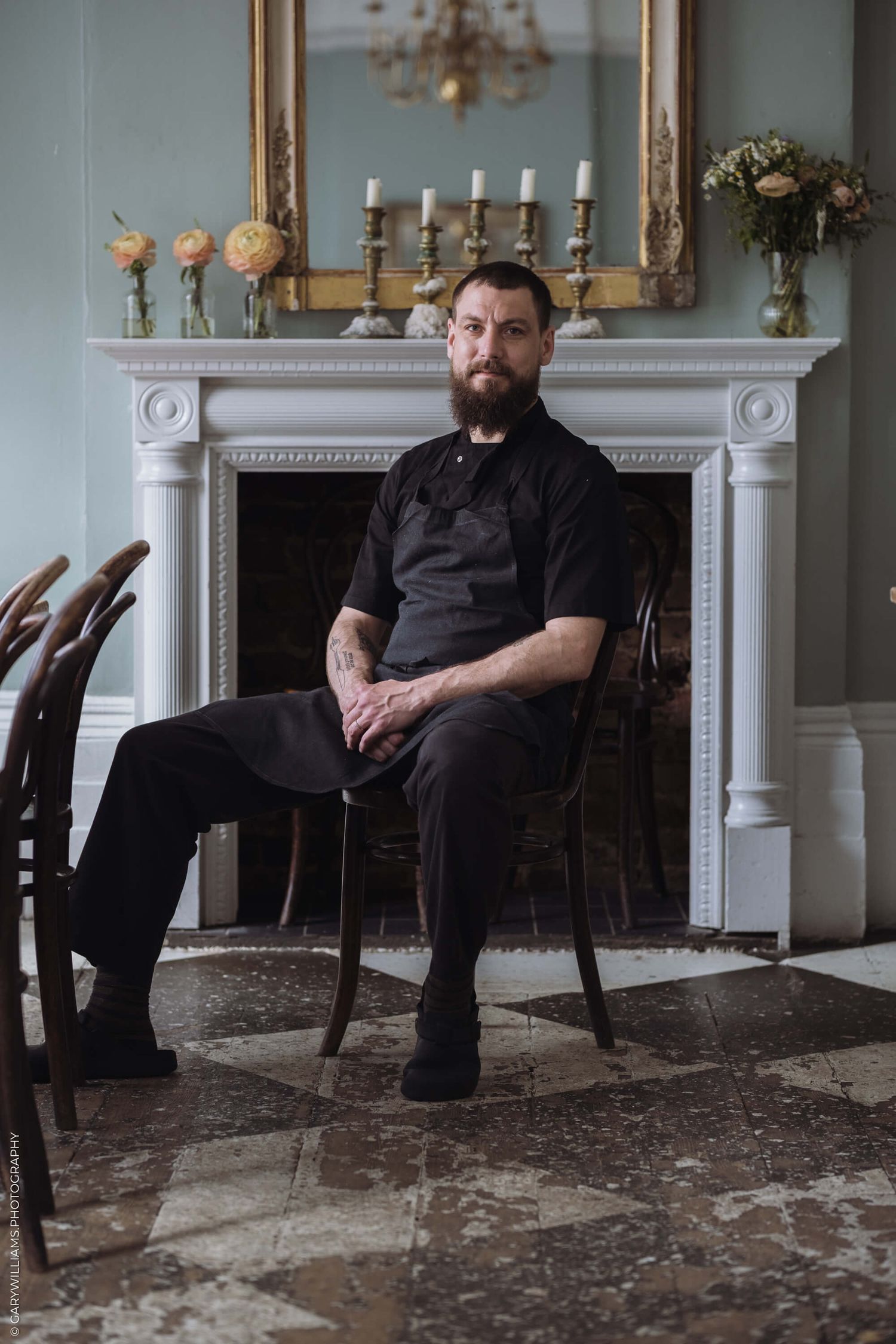 Someone wearing all black sits in an elegant room with a white fireplace mantel, candelabra and floral arrangements.