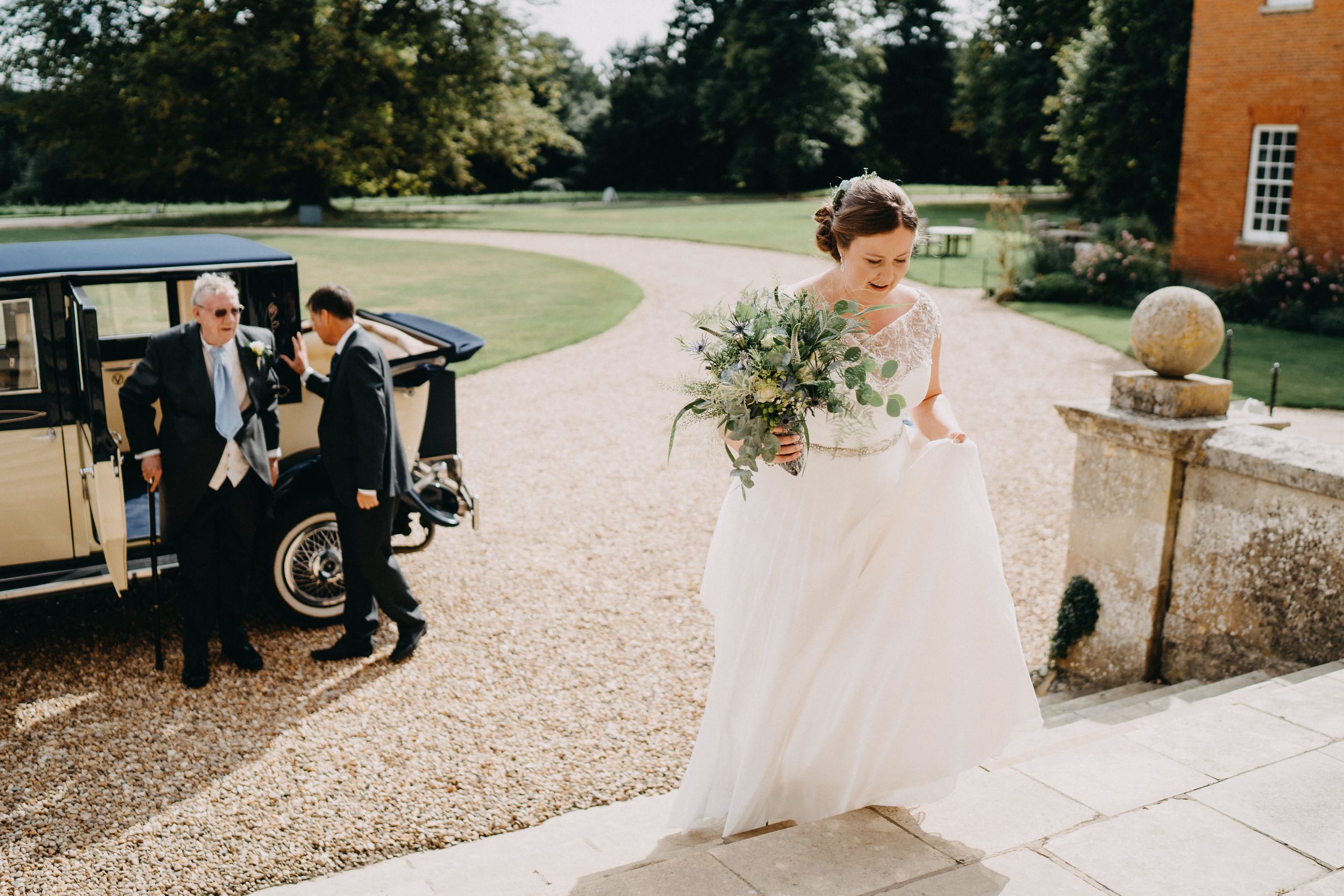A classic vintage car waits on a gravel driveway outside an elegant brick mansion during a formal event.