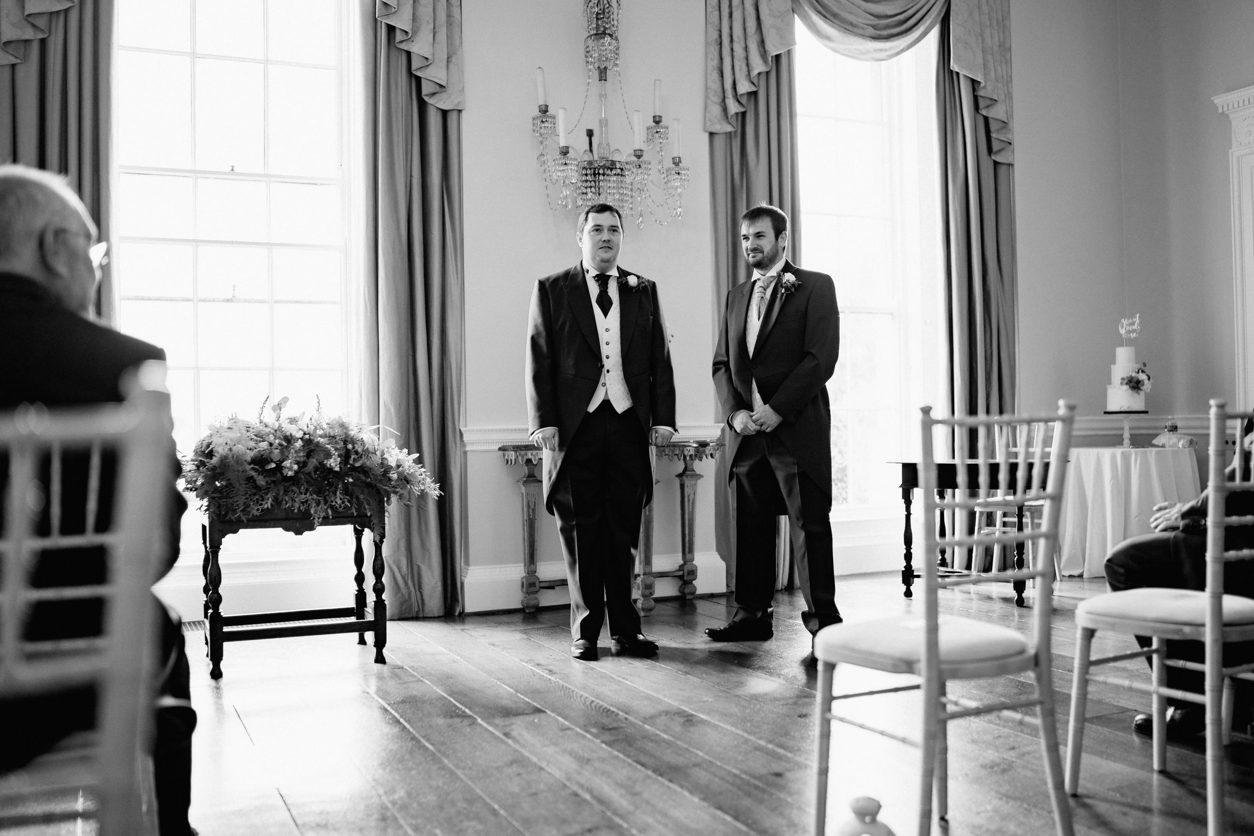 Two figures in formal attire stand in an elegant room with draped windows and wooden floors at Kelmarsh Hall.