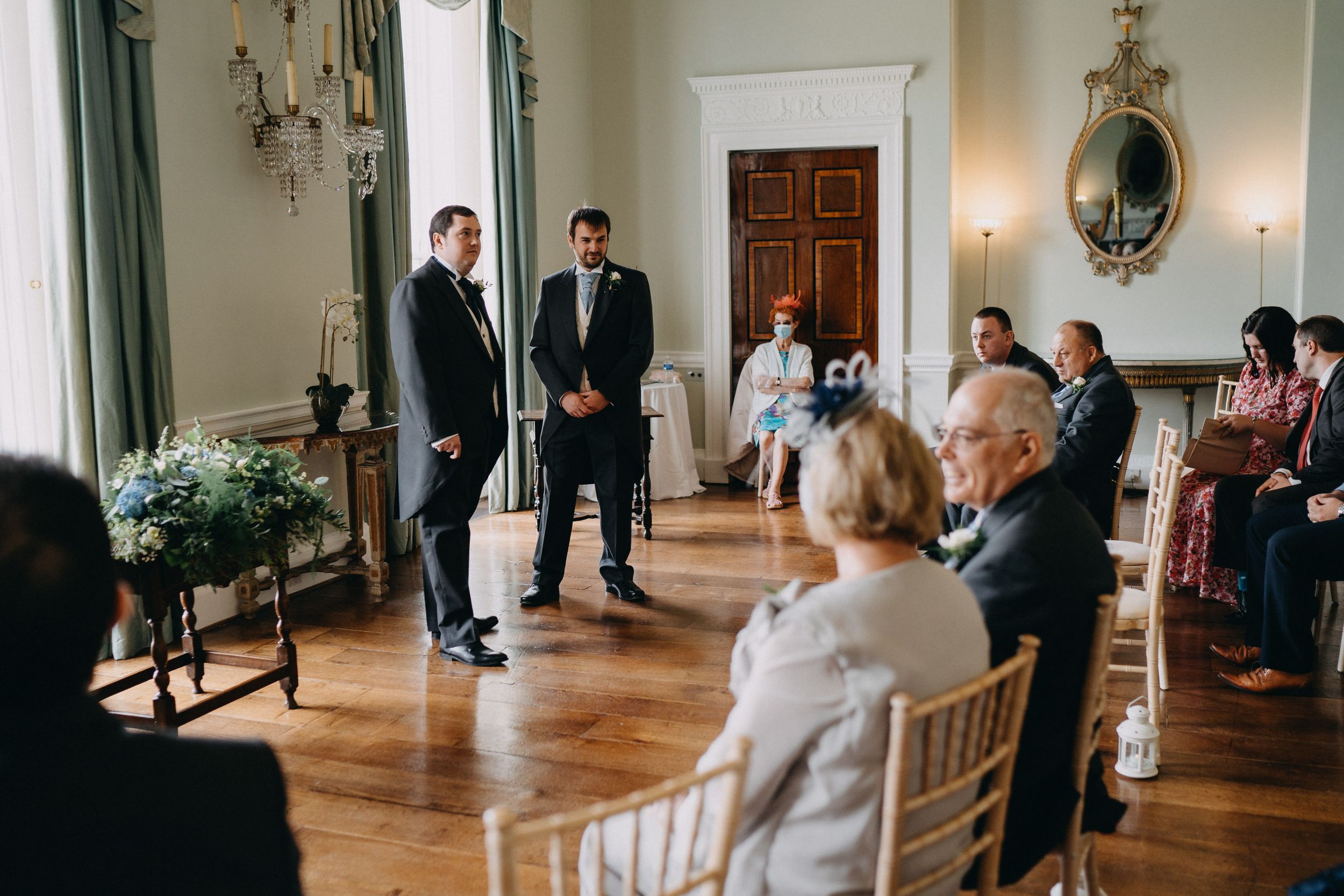 Guests seated in elegant chairs attend a formal indoor ceremony in a stately room of Kelmarsh Hall with chandeliers.