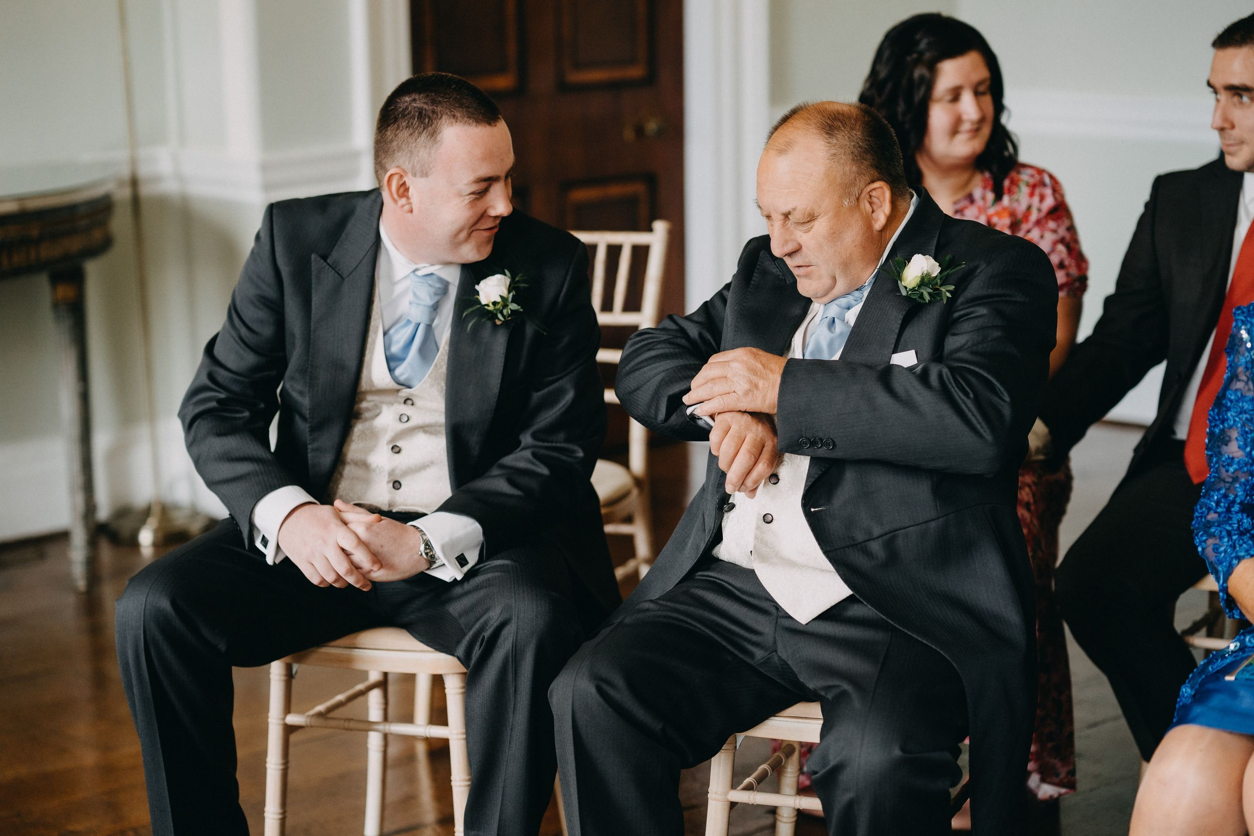 Two guests in formal attire share a conversation while seated on wooden chairs at an event.