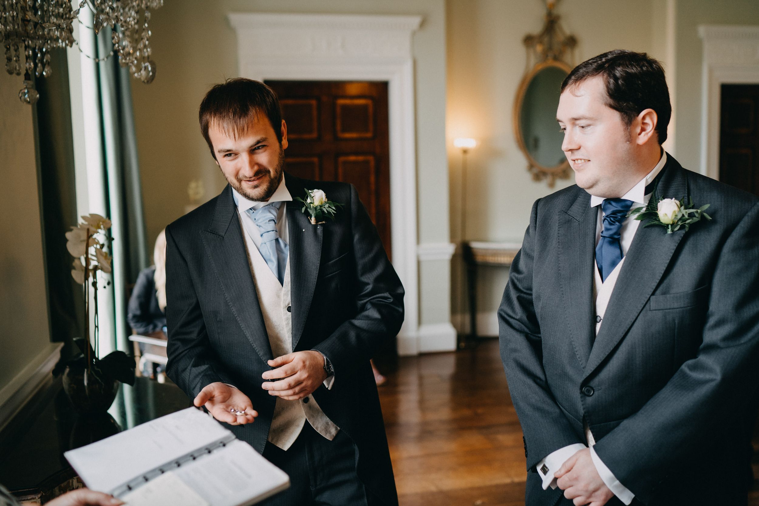 Two figures in dark suits stand near formal documents in an elegant room with hardwood floors.