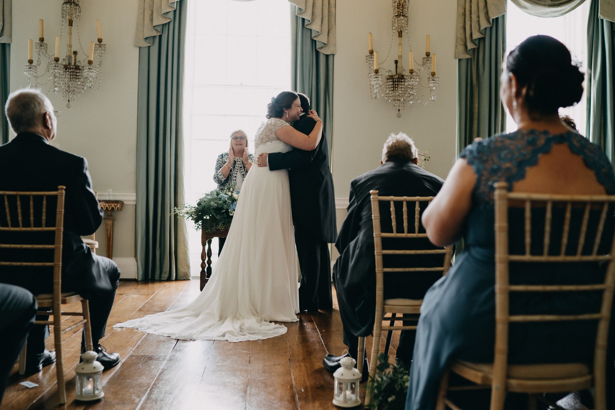 An intimate wedding ceremony at Kelmarsh Hall taking place in a classic room with chandelier lighting and gathered guests.