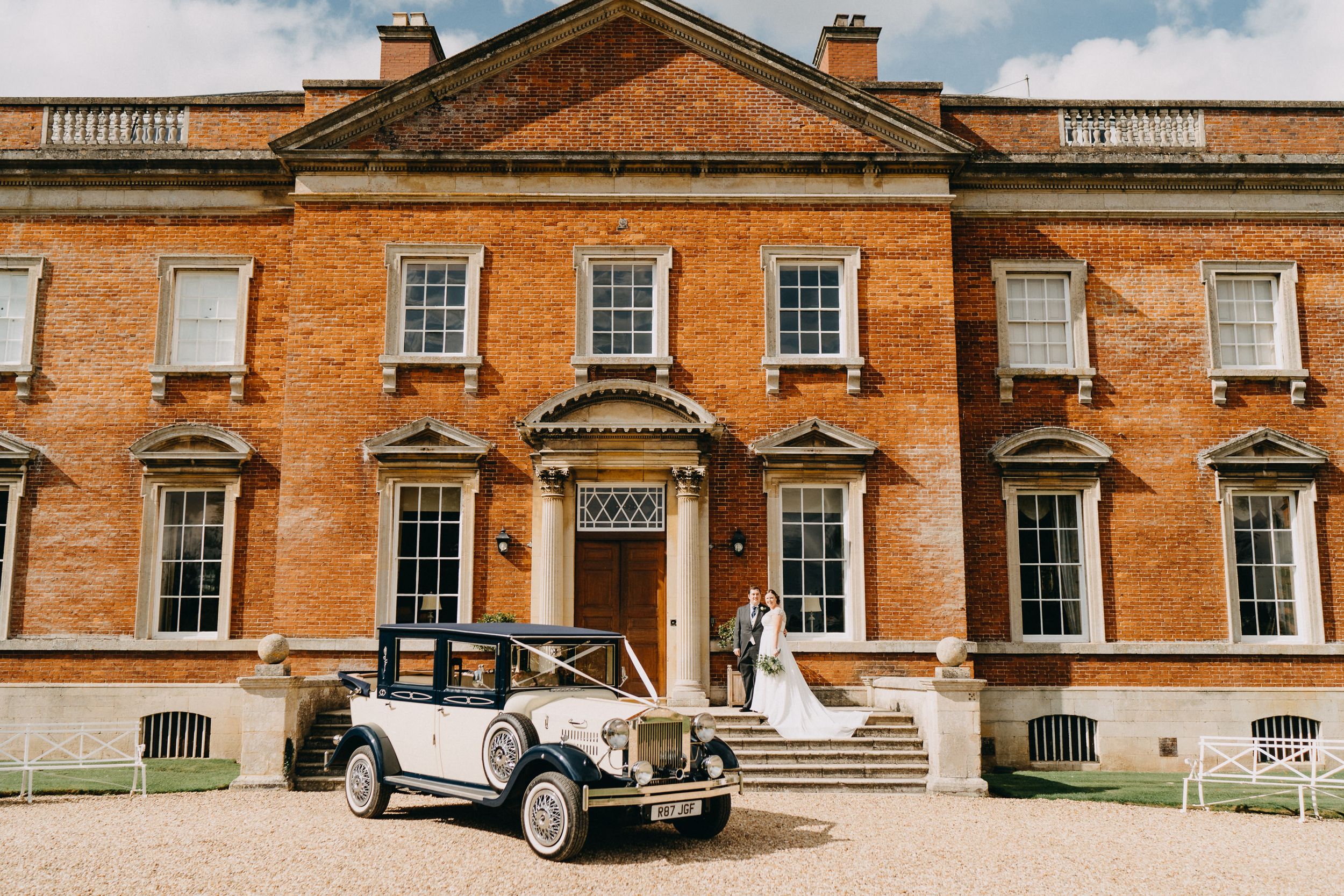 A grand Georgian-style mansion of Kelmarsh Hall with beautiful symmetrical architecture and a vintage car parked outside.