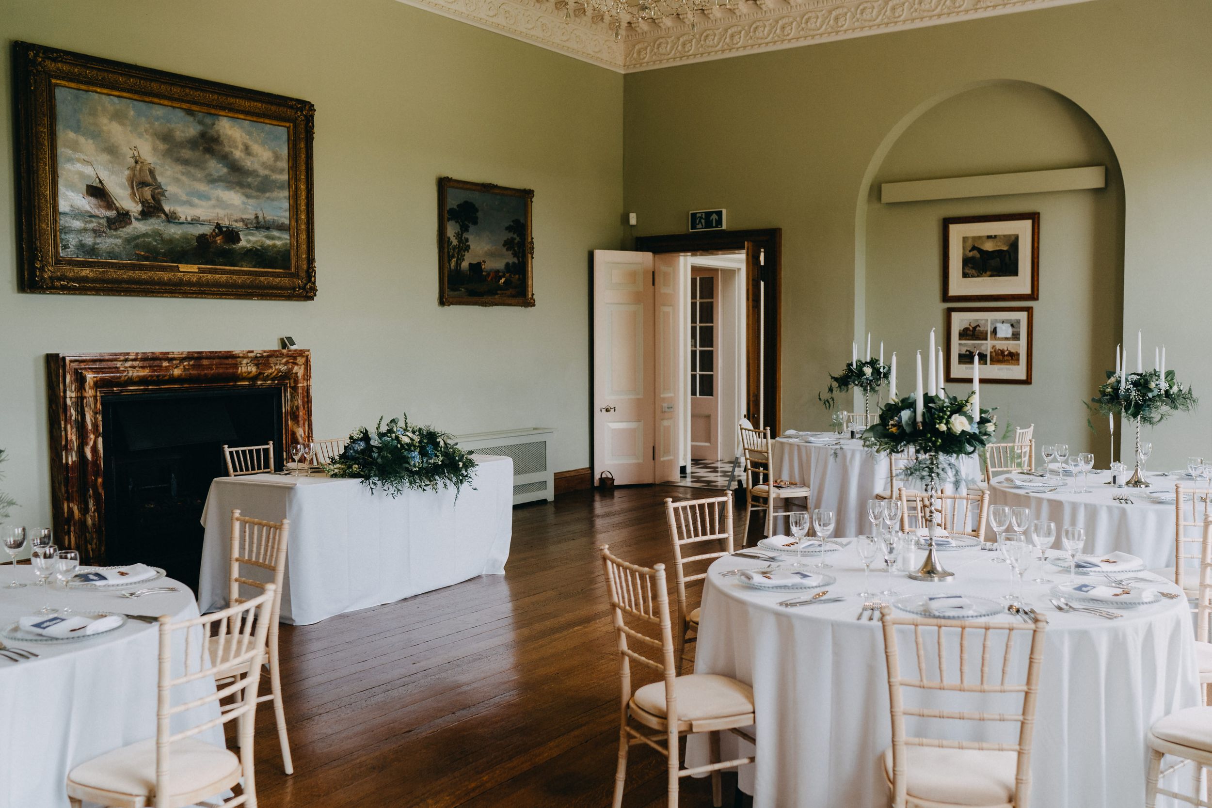 An elegant wedding reception room at Kelmarsh Hall with white tables, classic artwork, and traditional furnishings.