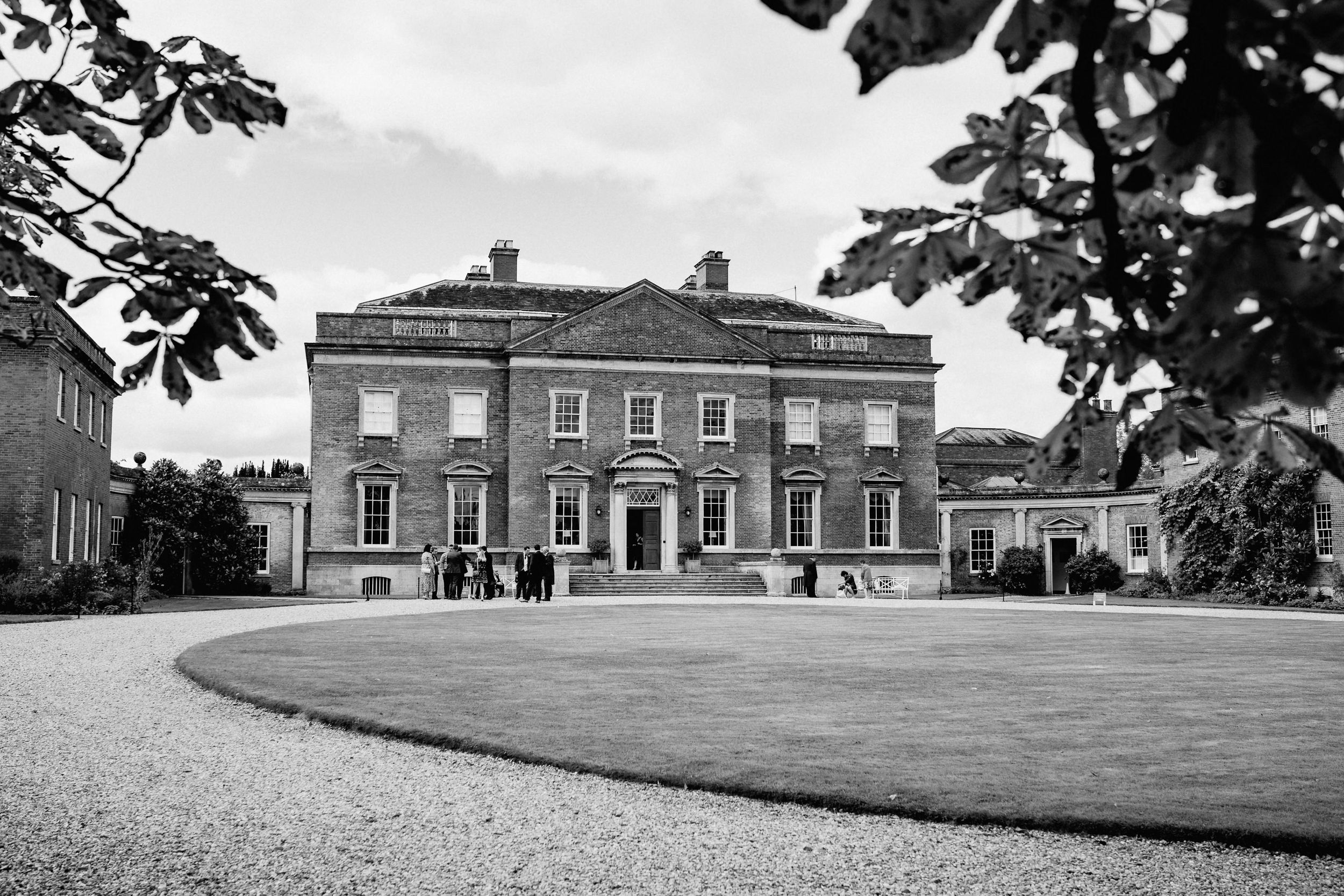 A black and white photograph of a stately Georgian manor house of Kelmarsh Hall with gravel driveway.