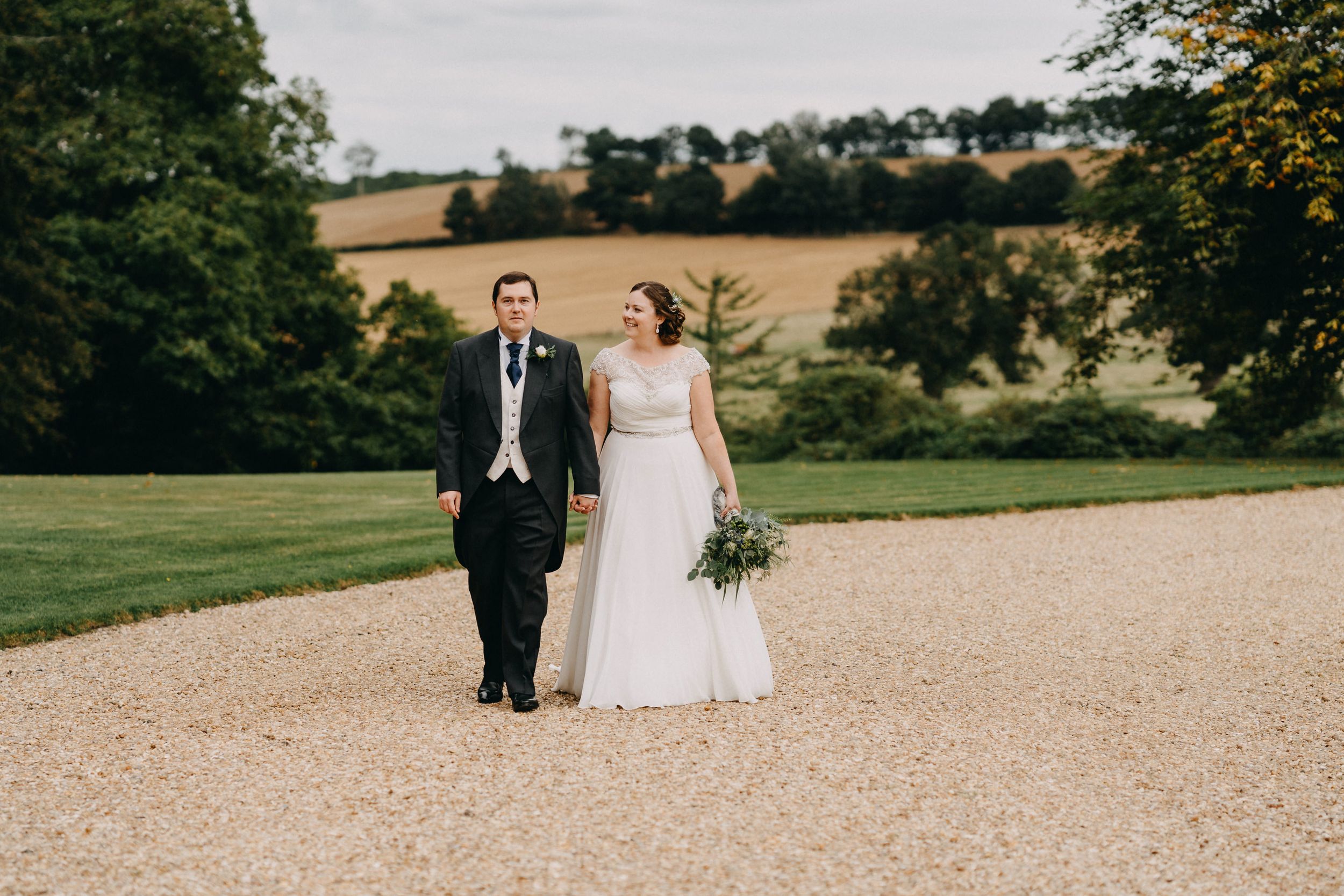 Couple walking together on a gravel path surrounded by green countryside on their wedding day.