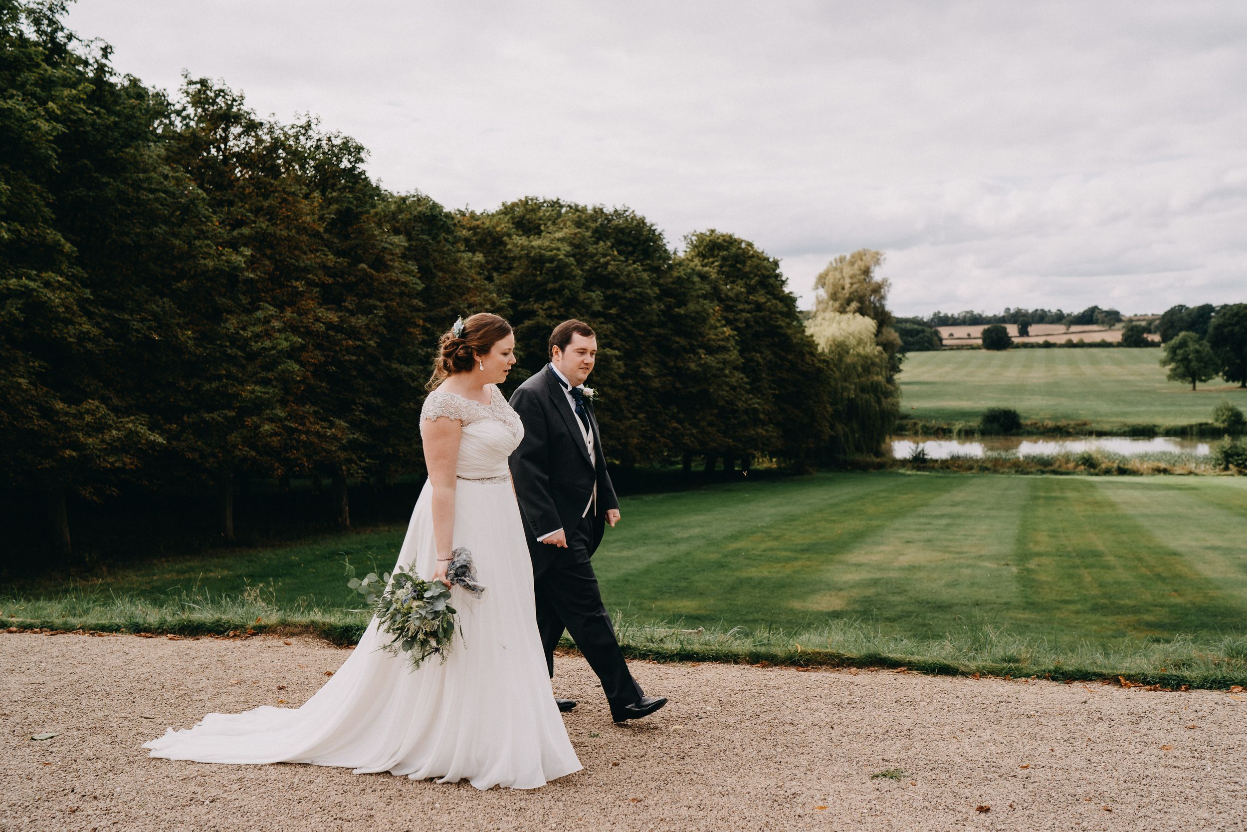 Couple strolling along a gravel path with scenic countryside views on their wedding day.
