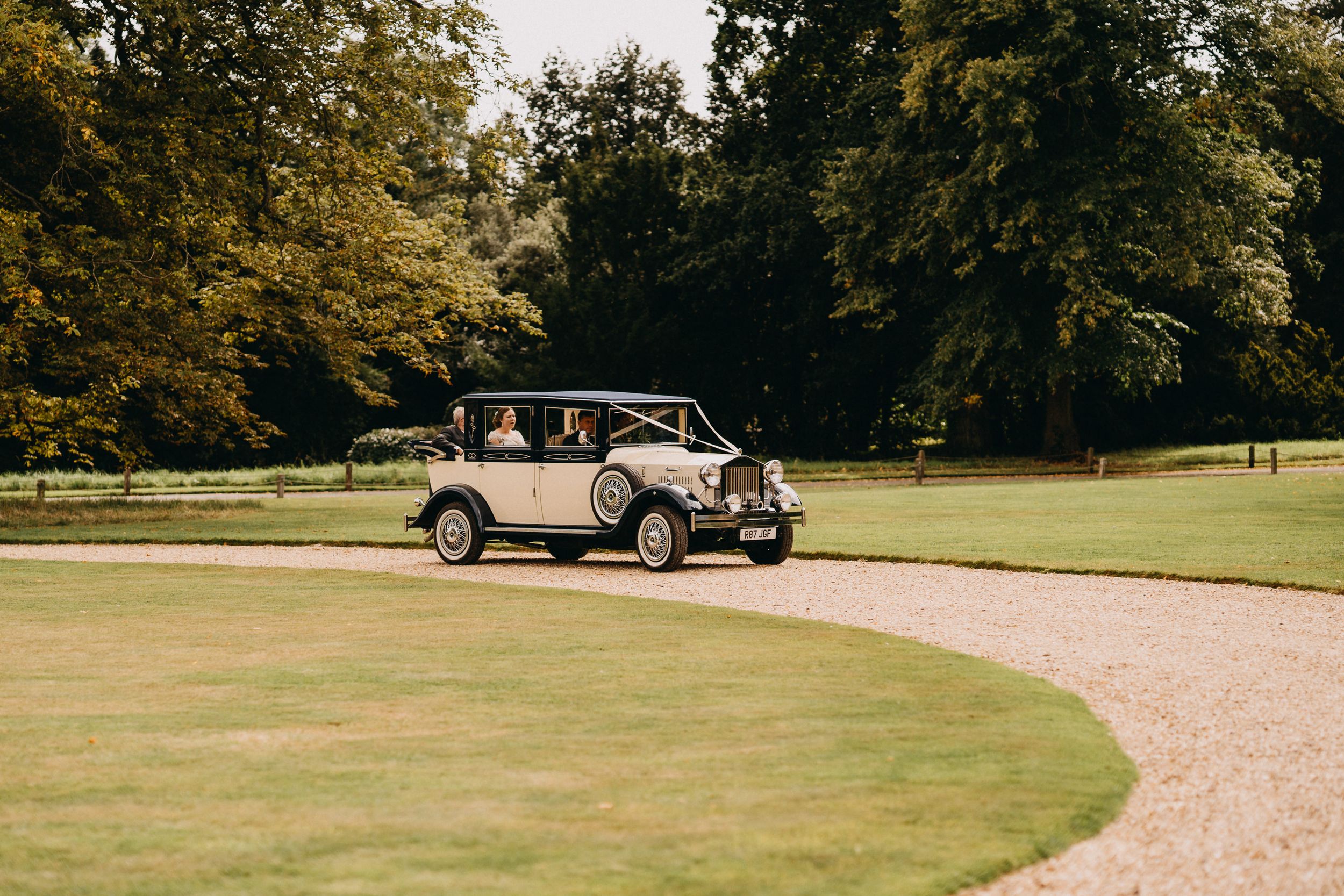 Vintage cream-colored wedding car parked on curved driveway surrounded by trees.